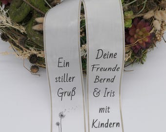 Puede incluir: Una cinta blanca con ribete dorado y flecos negros, con el texto "Ein stiller Gruß" y "Deine Freunde Bernd & Iris mit Kindern." Una pancarta verde arriba dice "Trauerschleifenmanufaktur.de Aus Tradition seit 1947."