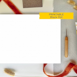 Puede incluir: Papel cuadriculado de puntos negros imprimible. El papel es blanco con puntos negros. El papel está rodeado de materiales de artesanía, incluyendo un carrete de cinta roja, un punzón de madera y un libro encuadernado en cuero marrón.
