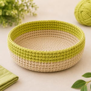 May include: A round, shallow crochet basket featuring a two-tone design with a beige base and a lime green upper rim. The basket is constructed with a visible, textured stitch pattern, creating a sturdy, circular container with a flat bottom.