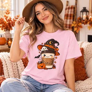 May include: A light pink short-sleeve t-shirt featuring a graphic print of a coffee cup wearing a witch hat with a small ghost, a pumpkin, and a bat. The design includes orange and black accents, stars, and autumn leaves, centred on the chest.