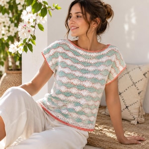 Puede incluir: Una blusa de ganchillo de manga corta con un patrón horizontal ondulado en rayas alternas de color blanco, verde claro y melocotón. La prenda tiene un escote ancho y redondeado, y está rematada con un borde liso de color melocotón.