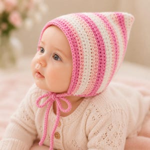 May include: A crochet bonnet featuring horizontal stripes in shades of pink, light pink, and white. The pointed hat has a textured knit pattern and includes two matching pink yarn ties that fasten under the chin with a simple bow knot.