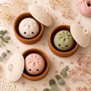 May include: Three crocheted stuffed toys shaped like steamed buns, each resting in a brown circular basket with a matching off-white lid. The buns are white, light green, and light pink, featuring embroidered black eyes, smiles, and pink cheeks.