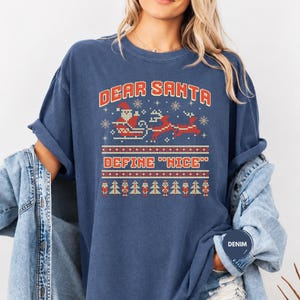 May include: A blue short-sleeve t-shirt featuring a red and white graphic print with the text "DEAR SANTA DEFINE "NICE"" above a pixelated illustration of Santa in a sleigh with reindeer, snowflakes, and a row of nutcrackers and Christmas trees.