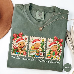 May include: A moss green short-sleeve t-shirt featuring three postage stamp-style illustrations of gingerbread people wearing Santa hats. The text below reads 'tis the season to reinforce behavior'. The shirt has a crew neckline and a Comfort Colors label.