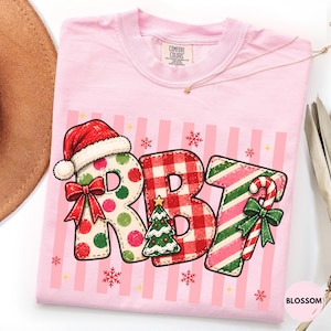 May include: A light pink t-shirt featuring a graphic of the letters RBT. The R has a red Santa hat and a red bow. The B features a small Christmas tree. The T is decorated with a candy cane and a green bow. The letters are set against a pink striped background with small red snowflakes.