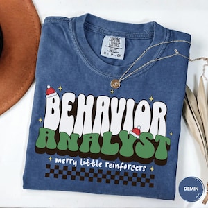 May include: A blue short-sleeve t-shirt with a graphic print that reads BEHAVIOR ANALYST in white and green letters, topped with small Santa hats. Below, the text reads merry little reinforcers above a black and white checkered pattern.