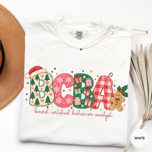 May include: A white t-shirt featuring the text BCBA and board certified behavior analyst in red script. The letters are decorated with Christmas trees, snowflakes, gift boxes, and a red plaid pattern, topped with a Santa hat and gingerbread man.