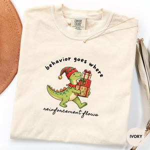 May include: An ivory t-shirt featuring a graphic of a green dinosaur wearing a red and green elf hat and collar, holding a stack of three wrapped Christmas gifts. The text "behavior goes where reinforcement flows" is printed in black lettering.