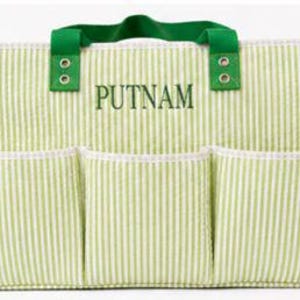 May include: A green and white striped tote bag with three front pockets and a green handle. The word "PUTNAM" is embroidered on the front. A small potted plant is to the left of the bag.