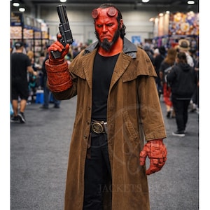 May include: A person dressed as Hellboy, holding a gun. They wear a long brown coat, black pants, and boots. Their skin is red, with horns and a beard. The costume includes a large red glove on the right hand.