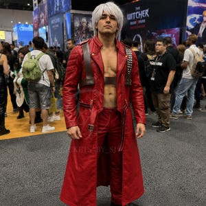 May include: A person in a red leather cosplay outfit, including a long coat, pants, and a shoulder strap. The coat has silver studded accents. The person has white hair and is standing in a crowded convention hall.