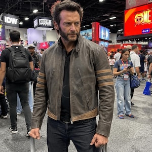 May include: A man wearing a brown leather jacket with tan stripes on the sleeves and dark blue jeans. The man is at a convention with other people in the background. The man has a beard and is holding metal claws.