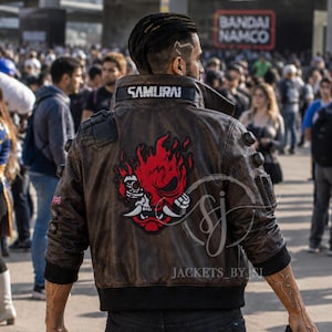 May include: A brown leather jacket featuring a large red and black graphic of a stylized oni mask on the back. The collar has the word SAMURAI printed in white. The jacket includes black ribbed cuffs and hem, with small circular studs on the shoulders.