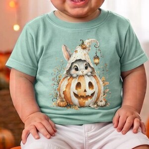 May include: A light green short-sleeved t-shirt featuring a graphic print of a small white rabbit wearing a pointed hat and sitting inside a carved orange jack-o'-lantern pumpkin, paired with plain white shorts.