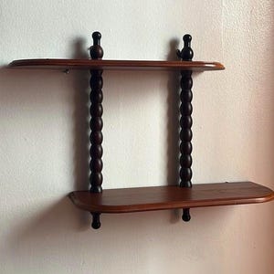 以下が含まれることがあります： 濃い茶色の仕上げが施された、2段の木製ウォールシェルフ。棚は旋盤加工されたスピンドルで支えられ、装飾的なフィニアルで飾られています。白い壁に取り付けられた棚です。