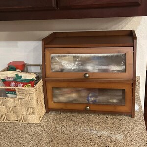 May include: A two-tiered wooden bread bin with glass panel doors. The bread bin is made of light brown wood and has a dark brown top. The glass panels have a ribbed texture. The bread bin has two small brass knobs for opening the doors.