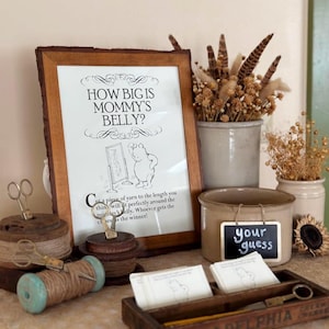 May include: A framed Winnie the Pooh themed game with the text "HOW BIG IS MOMMY'S BELLY?" is displayed. The scene includes spools of thread, scissors, a small chalkboard sign that says "your guess", and a wooden box with the word "PHILADELPHIA" on it.