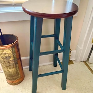 May include: A teal-painted wooden bar stool with a round, light-colored seat and a dark brown edge. The stool has three horizontal supports and is positioned near a window with blinds. A copper-colored fire extinguisher is visible in the background.