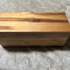 May include: A rectangular wooden box with a lid. The box is made of light brown wood with visible grain patterns and knots. The lid fits snugly on top, creating a clean, minimalist design. The box appears to be new and unused.