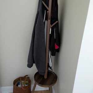 May include: A brown wooden coat stand with multiple branches for hanging clothes. A dark grey hoodie and a black jacket hang on the stand. A brown leather tote bag sits on the floor beside the stand.