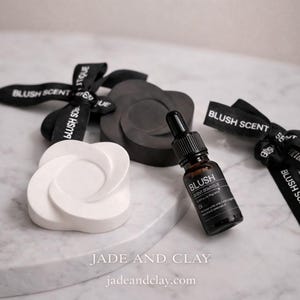 May include: A small brown glass bottle with a black dropper labeled "BLUSH" sits next to a white and a black decorative object. Black ribbons with "BLUSH SCENT" text are also present. The website "jadeandclay.com" is visible.