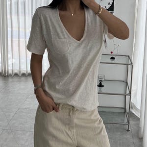 May include: A cream-colored V-neck t-shirt with a small pocket on the chest. The shirt has short sleeves and a relaxed fit. The model is wearing light beige pants. The overall style is casual and comfortable.