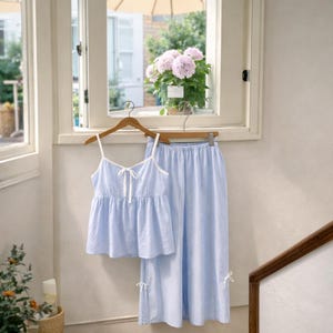 Puede incluir: Un conjunto de pijama azul claro con una camiseta de tirantes y pantalones a juego. La camiseta tiene ribetes blancos y un lazo. Los pantalones tienen un corte holgado y lazos en los tobillos. El conjunto est&aacute; colgado en una percha de madera.