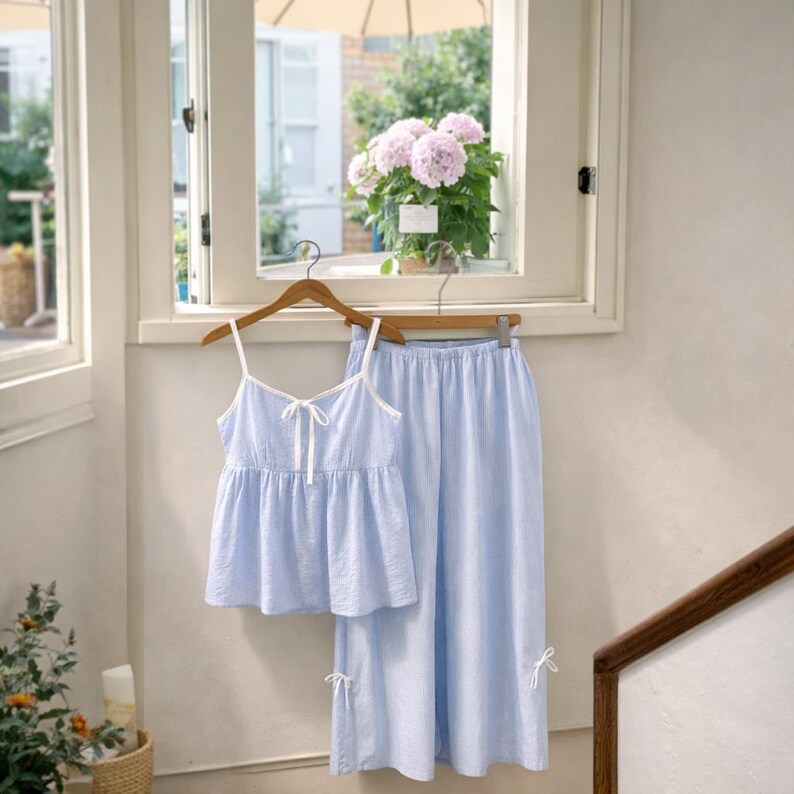 Puede incluir: Un conjunto de pijama azul claro con una camiseta de tirantes y pantalones a juego. La camiseta tiene ribetes blancos y un lazo. Los pantalones tienen un corte holgado y lazos en los tobillos. El conjunto est&aacute; colgado en una percha de madera.