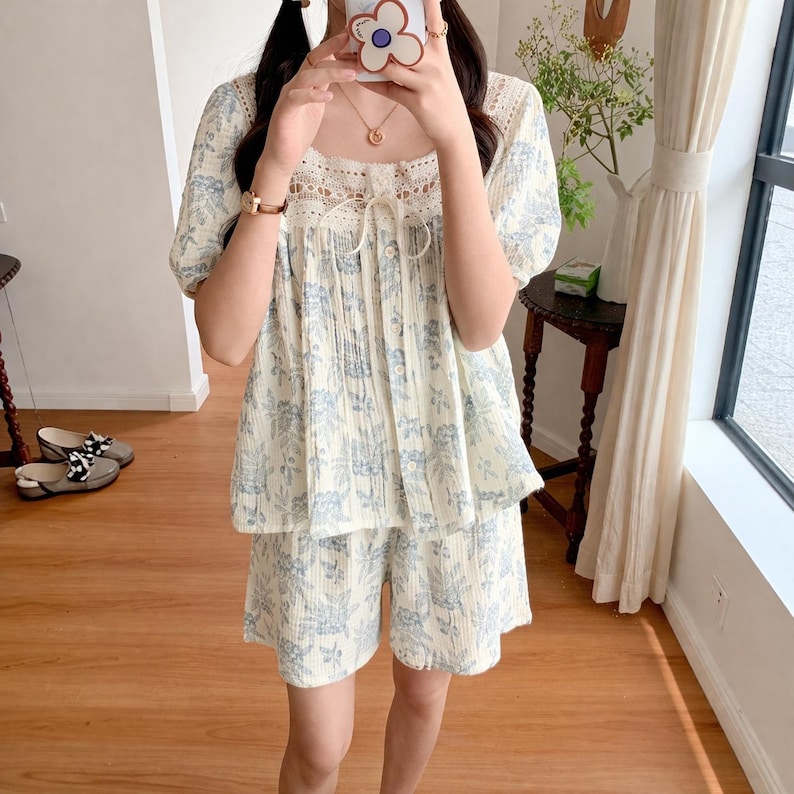 May include: A two-piece pajama set in a cream color with a blue floral pattern. The top has short sleeves and a square neckline with lace trim. The shorts have a matching floral design. The set is made of a lightweight fabric.