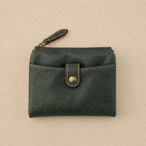 May include: Dark green leather wallet with a zippered top and a snap closure. The wallet has a small pocket on the front and white stitching details. The zipper pull is dark green leather with white stitching.