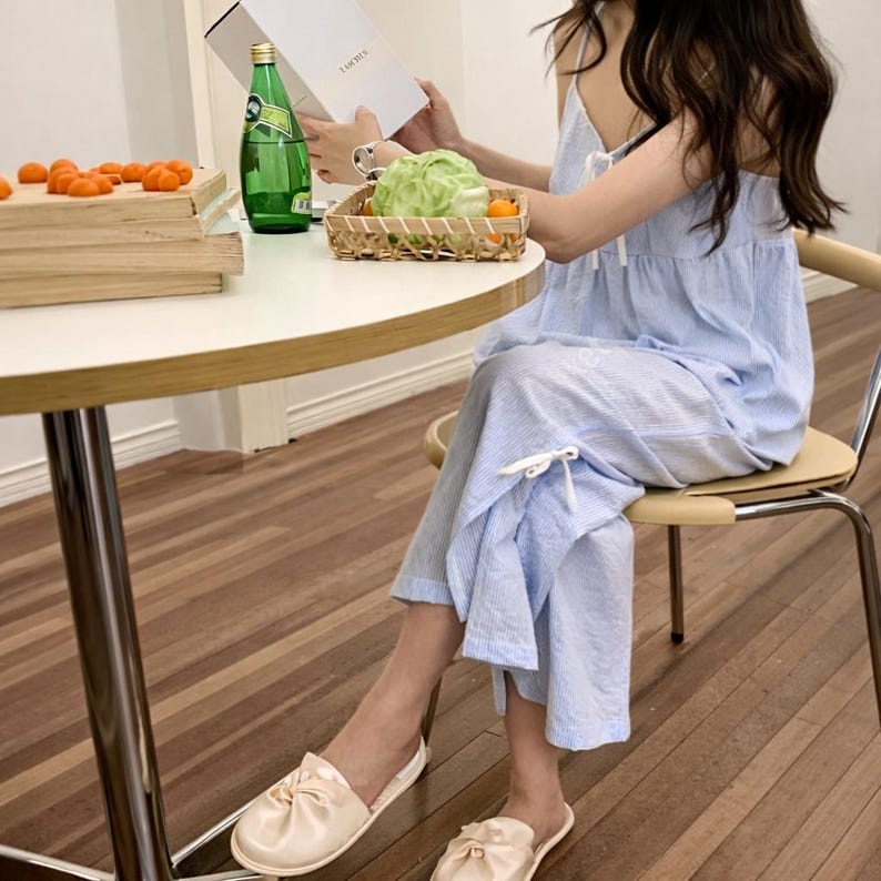 Puede incluir: Una persona con un conjunto de pijama a rayas azul claro con top y pantal&oacute;n a juego. La persona lleva zapatillas de color rosa claro. Una mesa redonda tiene una caja de madera con fruta naranja, una botella verde y una cesta con verduras verdes y naranjas.