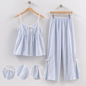 Puede incluir: Conjunto de pijama a rayas azul claro y blanco. El conjunto incluye una camiseta sin mangas con ribete blanco y detalle de lazo, y pantalones de pernera ancha a juego. Los pantalones tienen lazos en los tobillos. El conjunto de pijama est&aacute; colgado en una percha de madera.