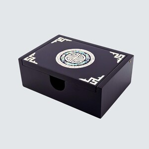 May include: A black rectangular box with a hinged lid. The lid features a circular inlaid design with mother-of-pearl and turquoise accents. Decorative white corner and edge details add contrast.