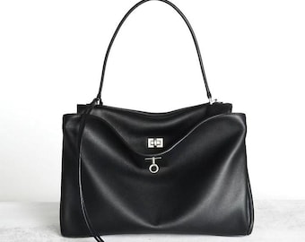 Puede incluir: Un bolso de mano de cuero negro con una sola asa arqueada, un cierre de giro rectangular de tono plateado y un dije circular plateado que cuelga de una fina correa de cuero en la parte frontal del bolso.