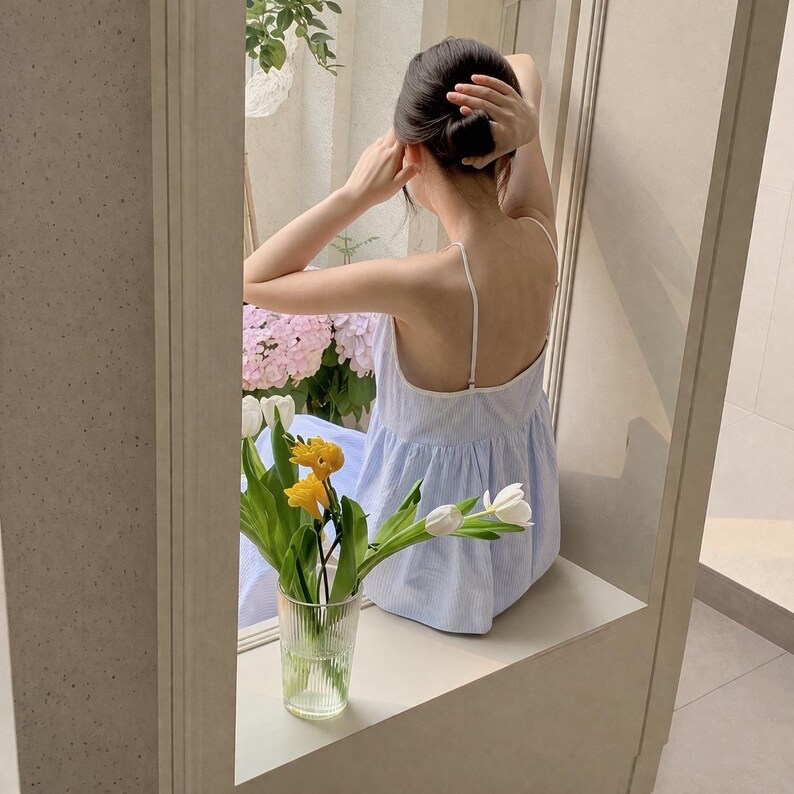 Puede incluir: Un vestido de verano a rayas azul claro y blanco con tirantes finos. Un jarr&oacute;n de cristal transparente contiene flores amarillas y blancas. La escena se refleja en un espejo, con flores rosas y vegetaci&oacute;n en el fondo.