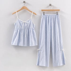 Puede incluir: Un conjunto de pijama a rayas azul claro y blanco. El conjunto incluye una camiseta sin mangas con lazo y tirantes finos, y pantalones de pernera ancha a juego. Los pantalones tienen lazos en los tobillos. El conjunto est&aacute; colgado en perchas de madera.