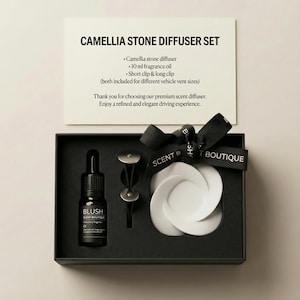 May include: A Camellia Stone Diffuser Set in a black box. The set includes a white camellia stone diffuser, a 10 ml bottle of fragrance oil, and clips for different vehicle vent sizes. A black ribbon with "SCENT BOUTIQUE" text is tied around the box.