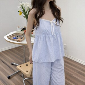 Puede incluir: Conjunto de pijama a rayas azules y blancas. La parte superior es de estilo camisola con lazo y ribete blancos. Los pantalones a juego son de pernera ancha. La tela parece ser una mezcla ligera de algod&oacute;n o lino.