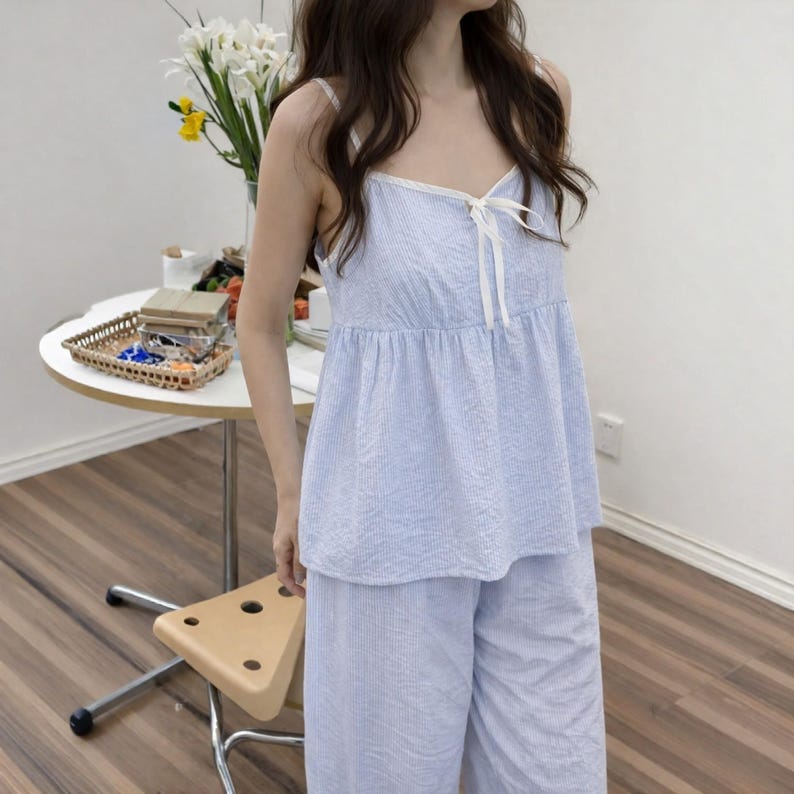 Puede incluir: Conjunto de pijama a rayas azules y blancas. La parte superior es de estilo camisola con lazo y ribete blancos. Los pantalones a juego son de pernera ancha. La tela parece ser una mezcla ligera de algod&oacute;n o lino.