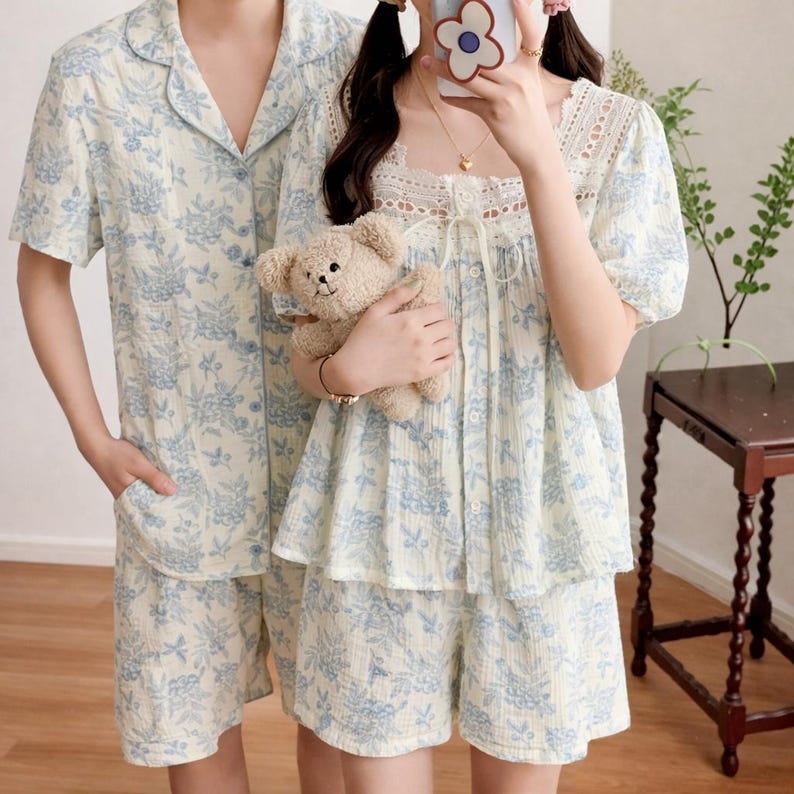 May include: Matching pajama sets in a light cream color with a blue floral pattern. The sets include a short-sleeved button-up shirt and shorts. One person is wearing a pajama set with a lace-trimmed top and holding a teddy bear.