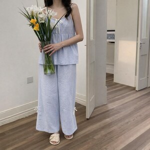 Puede incluir: Conjunto de pijama a rayas azul claro y blanco. El conjunto incluye un top de camisola con lazos y pantalones de pernera ancha. La persona sostiene un jarr&oacute;n de cristal transparente lleno de flores blancas y amarillas.