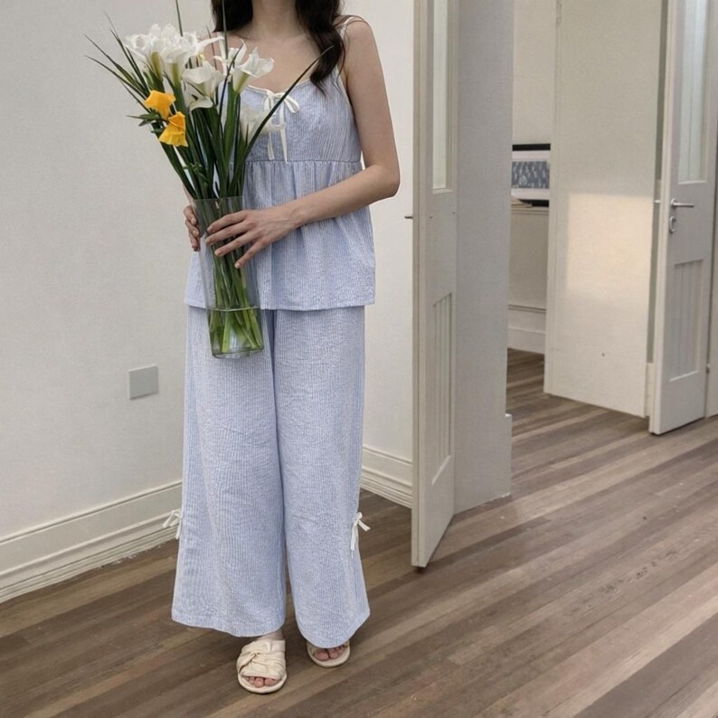 Puede incluir: Conjunto de pijama a rayas azul claro y blanco. El conjunto incluye un top de camisola con lazos y pantalones de pernera ancha. La persona sostiene un jarr&oacute;n de cristal transparente lleno de flores blancas y amarillas.