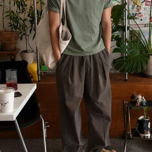 May include: A sage green short-sleeved crew neck t-shirt with rolled sleeves, paired with loose-fitting dark charcoal gray pleated trousers and a large off-white canvas tote bag carried over the shoulder, completed with beige thong sandals.