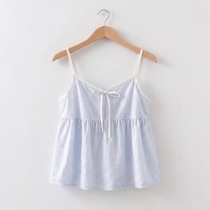 Puede incluir: Un top camisero a rayas azules y blancas con tirantes finos y un detalle de lazo en el escote. La parte superior est&aacute; colgada de una percha de madera sobre un fondo blanco. El camisero tiene la cintura fruncida.