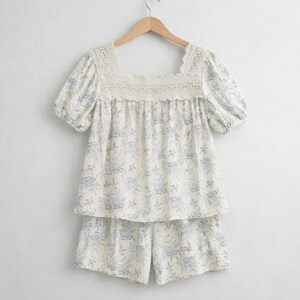 May include: A two-piece pajama set with a floral pattern. The top has a square neckline with lace trim and short sleeves. The shorts match the top's floral design. The set is cream-colored with blue floral accents.