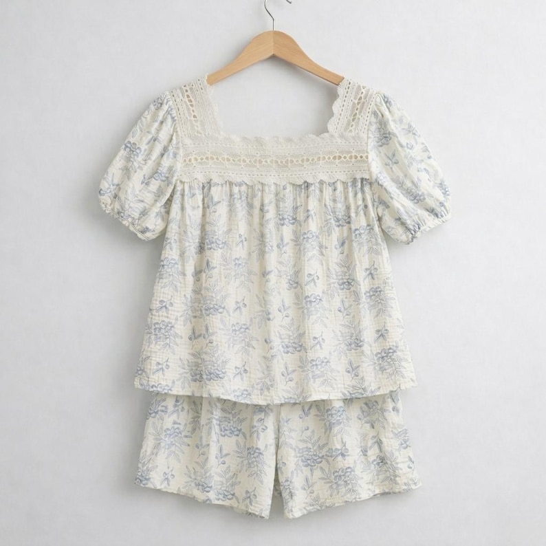 May include: A two-piece pajama set with a floral pattern. The top has a square neckline with lace trim and short sleeves. The shorts match the top's floral design. The set is cream-colored with blue floral accents.