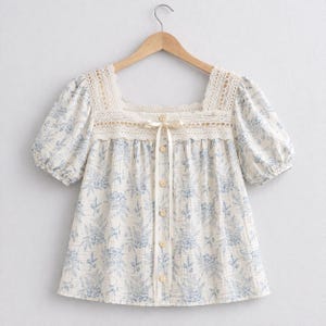 May include: A cream-colored blouse with a blue floral pattern and a square neckline. The blouse features short, puffy sleeves, a lace trim, and a row of buttons down the front. A cream-colored ribbon is tied at the neckline.