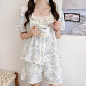 May include: A two-piece pajama set in white with a blue floral pattern. The top has a square neckline with lace trim and short, puffed sleeves. The matching shorts complete the set. A white mug with text is held in the image.