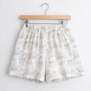 May include: Cream-colored shorts with a blue floral pattern hanging on a wooden hanger. The shorts have an elastic waistband and a loose fit. The fabric appears to be lightweight and breathable, suitable for warm weather.